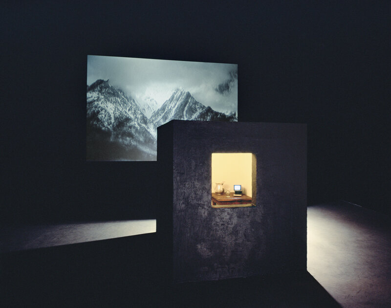 Bill Viola, Room for St. John of the Cross, 1983. Video and sound installation in a dark room, with a black cubicle with window, peat moss, wooden table, glass and metal pitcher with water, color video, and one-channel mono sound; black-and-white vi…